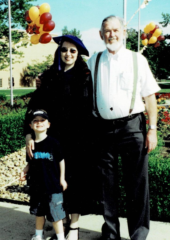 Outdoor family photo with red & gold balloons and ornate garden in background. Foreground shows older man on right, Janet in doctorate cap and robes and small 5 year old to her left. 