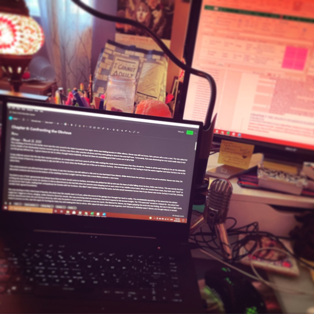 Blurred out photo of a black laptop with typed chapter and a big monitor int he background with a blurred outline for my book.